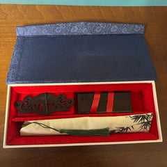 The opened fan box contains a fan stand and a fan pouch. The fan cover is painted with a bamboo leaf pattern, and a green tassel extends from the fan pouch.