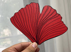 A hand holding a red gingko leaf shape fan panel in front of a white curtain