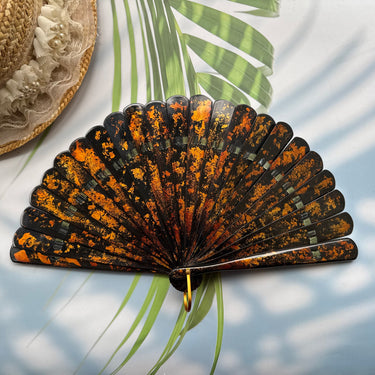 A Chinese lacquer folding fan with a gold-sprinkled pattern is placed on a table with sunlight, palm leaves and a straw hat in the background.