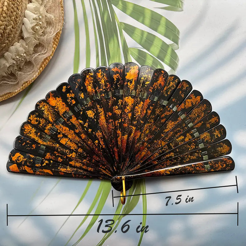 A Chinese lacquer folding fan with a gold-sprinkled pattern is placed on a table with sunlight, palm leaves and a straw hat in the background, with the size number indicated in the picture.