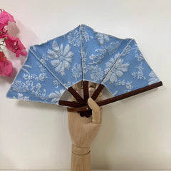 Classical embroidered hand fan displayed on a wooden hand, with delicate pink flower background.
