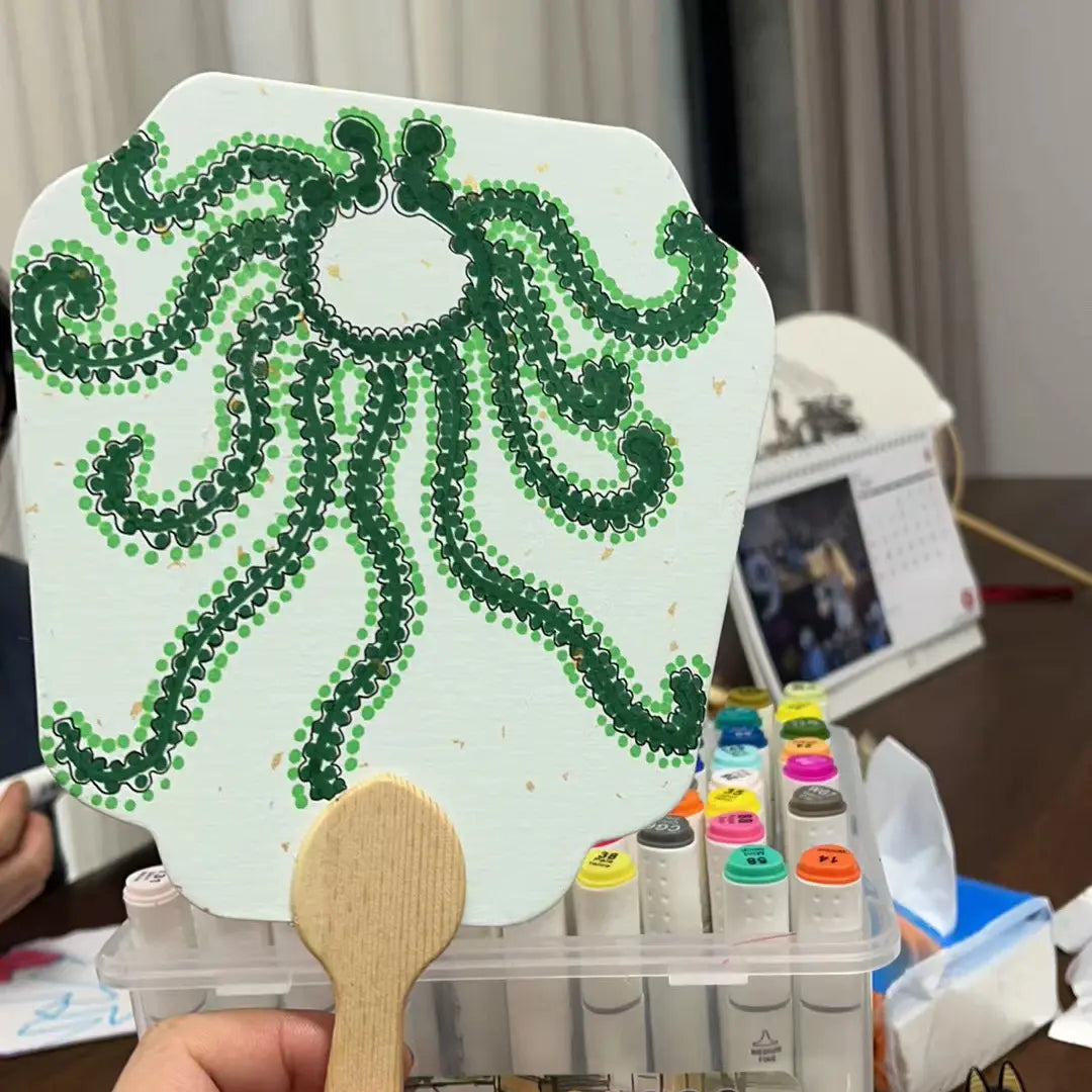 A hand holds a blank fixed fan made of gold-sprinkled paper, featuring a detailed octopus illustration. In the background, additional fans and a box of colored pens, a calendar and a bag of tissues are visible.