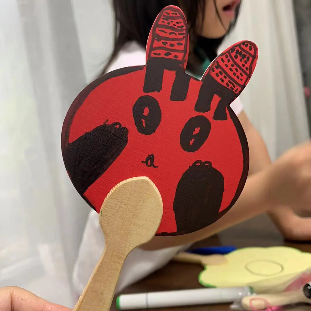 A hand holds a round fixed fan, featuring a child's drawing of a cute rabbit. Additional fans and a little girl are visible in the background.