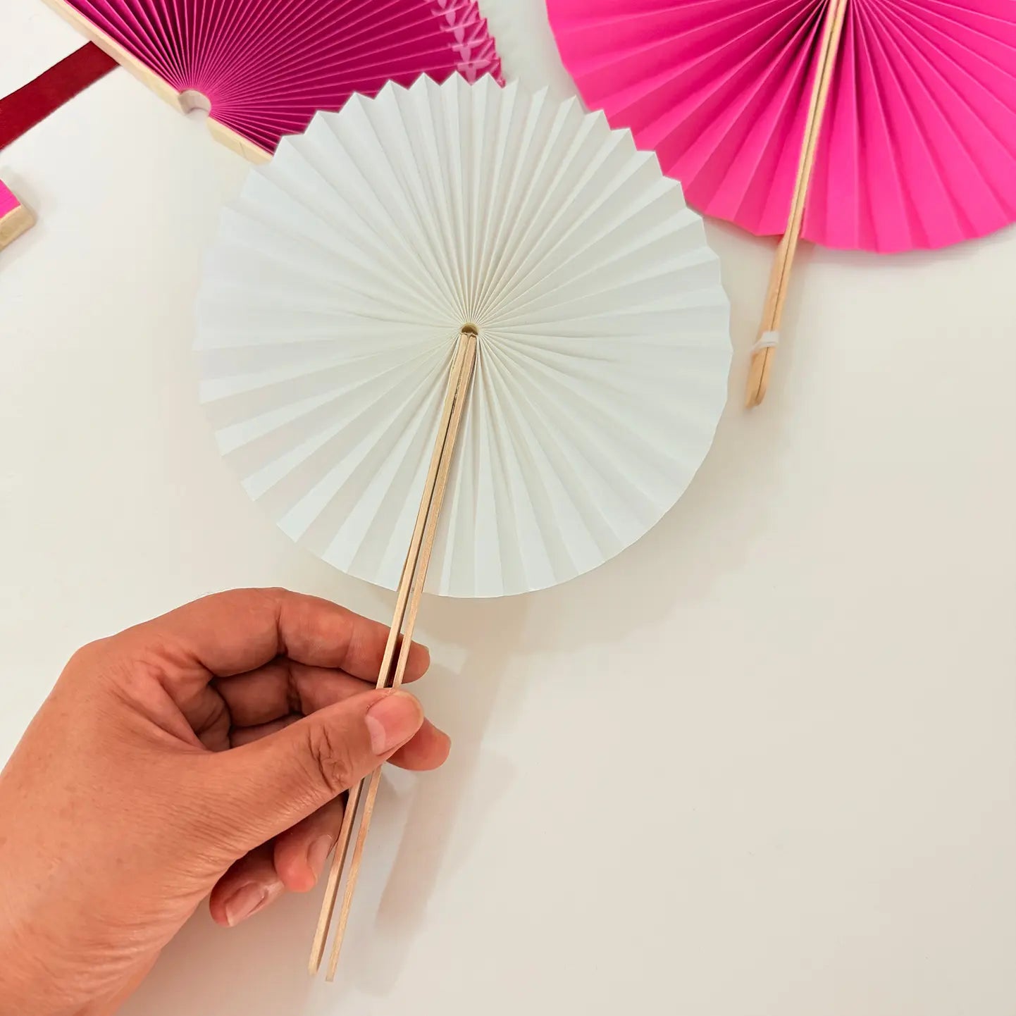 A hand holds a white round paper folding fan, with a colorful assortment of other round folding fans featuring diverse designs displayed in the background.