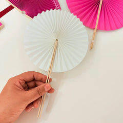 A hand holds a white round paper folding fan, with a colorful assortment of other round folding fans featuring diverse designs displayed in the background.
