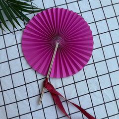 A rose-red round folding fan lies open on a textured cloth, secured in its unfolded position by a matching silk ribbon tied around the frame. Delicate green leaves are visible in the background.