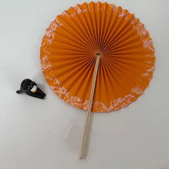 An orange round folding fan lies open on a white surface, secured in its unfolded position. Delicate chinese style patterns are printed on the outer layer of the fan, complemented by a lazy cat figurine decoration in the background.