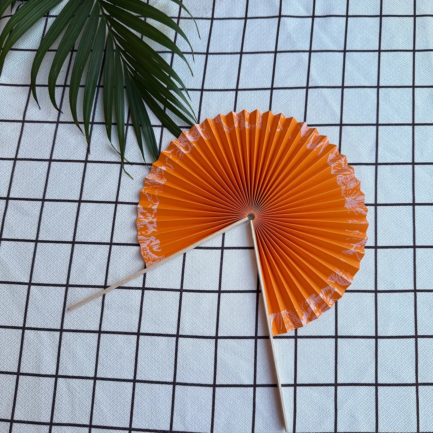 A orange round folding fan lies open on a textured cloth, placed in its half unfolded position. Delicate green leaves are visible in the background.
