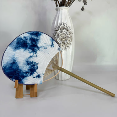A Japanese round fan with ice flower blue tie-dye print on the fan stand, with a white background and a vase
