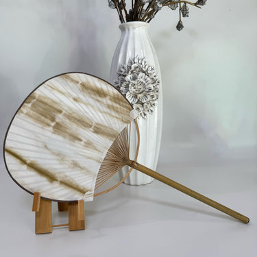 A Japanese round fan with bamboo brown tie-dye print on the fan stand, with a white background and a vase