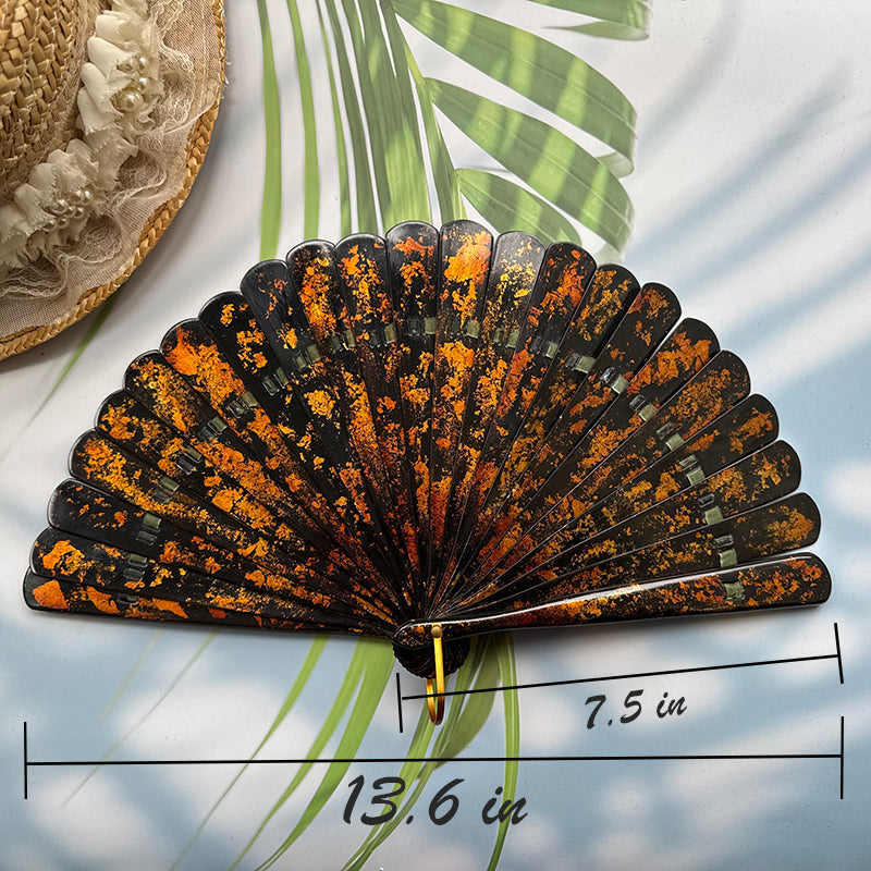 A Chinese lacquer folding fan with a gold-sprinkled pattern is placed on a table with sunlight, palm leaves and a straw hat in the background, with the size number indicated in the picture.