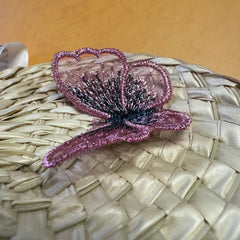 A delicate, shimmering pink and purple mesh butterfly with black detailing rests on a woven palm leaf fan. The fan’s natural texture and soft orange background create a warm, harmonious, and visually appealing composition, ideal for fashion or decor inspiration.