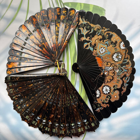 3 Chinese lacquer folding fan with gold-sprinkled patterns are placed on a table with sunlight and palm leaves in the background.
