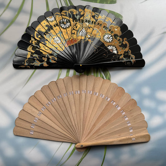 Two fans are placed on the table, one is a Chinese lacquer folding fan with intricate gold patterns, and the other is a wooden folding fan without lacquer or painting. The background has sun shadows, palm leaves and straw hat.