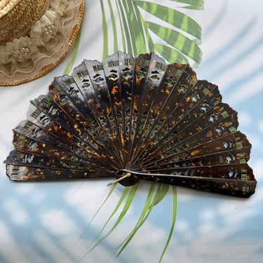 A Chinese lacquer folding fan (urushi fan) is placed on the table. The fan pattern is sprinkled with gold and has hollow carvings. The background has sunlight and shadows, palm leaves and straw hats.