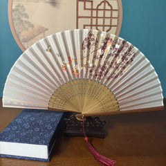 Chinese style hand held bamboo folding fan on a fan stand with handcrafted box on the side. The background is Chinese ink-wash window paper-cuts.