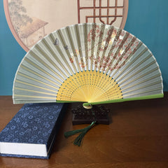 Chinese style hand held bamboo folding fan on a fan stand with handcrafted box on the side. The background is Chinese ink-wash window paper-cuts.