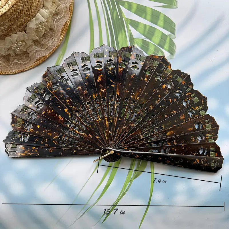 On the table lies an East Asian Lacquer Folding Fan (Urushi fan) with a hollowed-out rib design and gold-sprinkled patterns. The background is sunlight, palm leaves, and a straw hat. The dimensions of the fan are as shown in the picture.