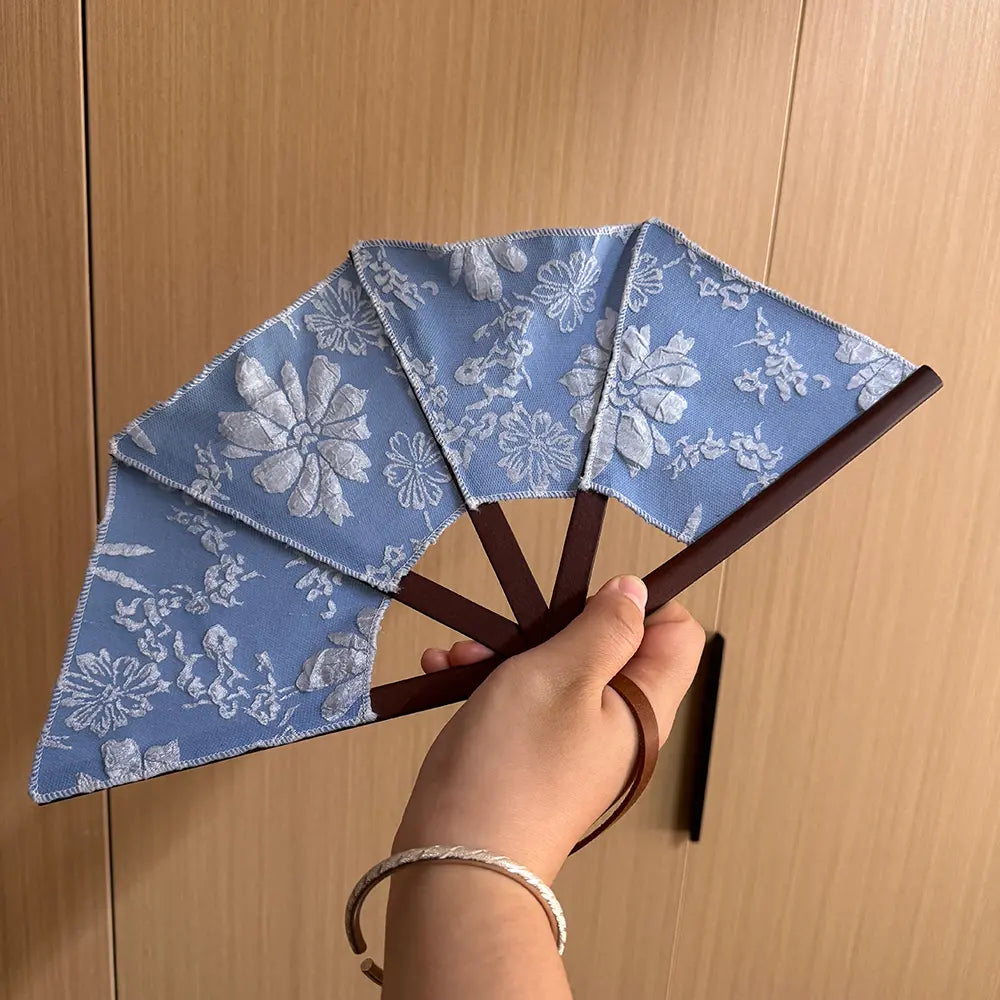 Blue classical embroidery hand fan held in hand, with its leather wrist strap wrapped around the wearer's wrist.