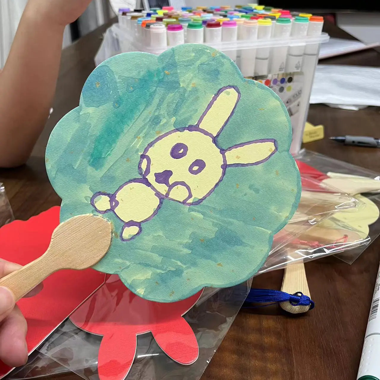 A hand holds a round fixed fan made of gold-sprinkled paper, featuring a child's drawing of a cute rabbit. Additional fans and a box of colored pens are visible in the background.