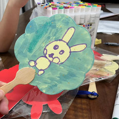 A hand holds a round fixed fan made of gold-sprinkled paper, featuring a child's drawing of a cute rabbit. Additional fans and a box of colored pens are visible in the background.