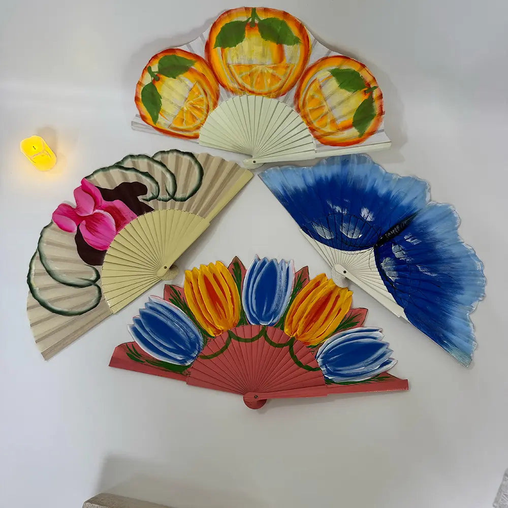 A styled still life flat lay of four beautiful Spanish hand fans. Each fan is uniquely hand-painted with vibrant designs: juicy orange slices, a blooming pink flower, a vivid blue butterfly, and cheerful yellow and blue tulips. Arranged on a clean white background with soft candlelight, creating an inviting Spanish-themed aesthetic.