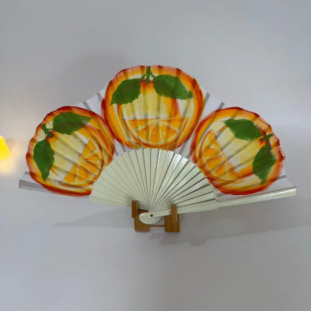 Decorative hand-painted Spanish fan featuring a juicy orange slices drawing. Styled shot on a white surface with a fan stand and a candle, evoking a Spanish ambiance.