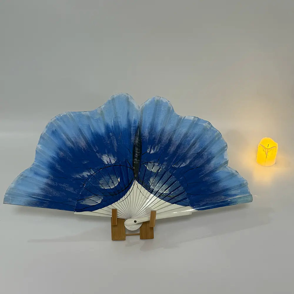 Decorative hand-painted Spanish fan featuring a vivid blue butterfly drawing. Styled shot on a white surface with a fan stand and a candle, evoking a Spanish ambiance.