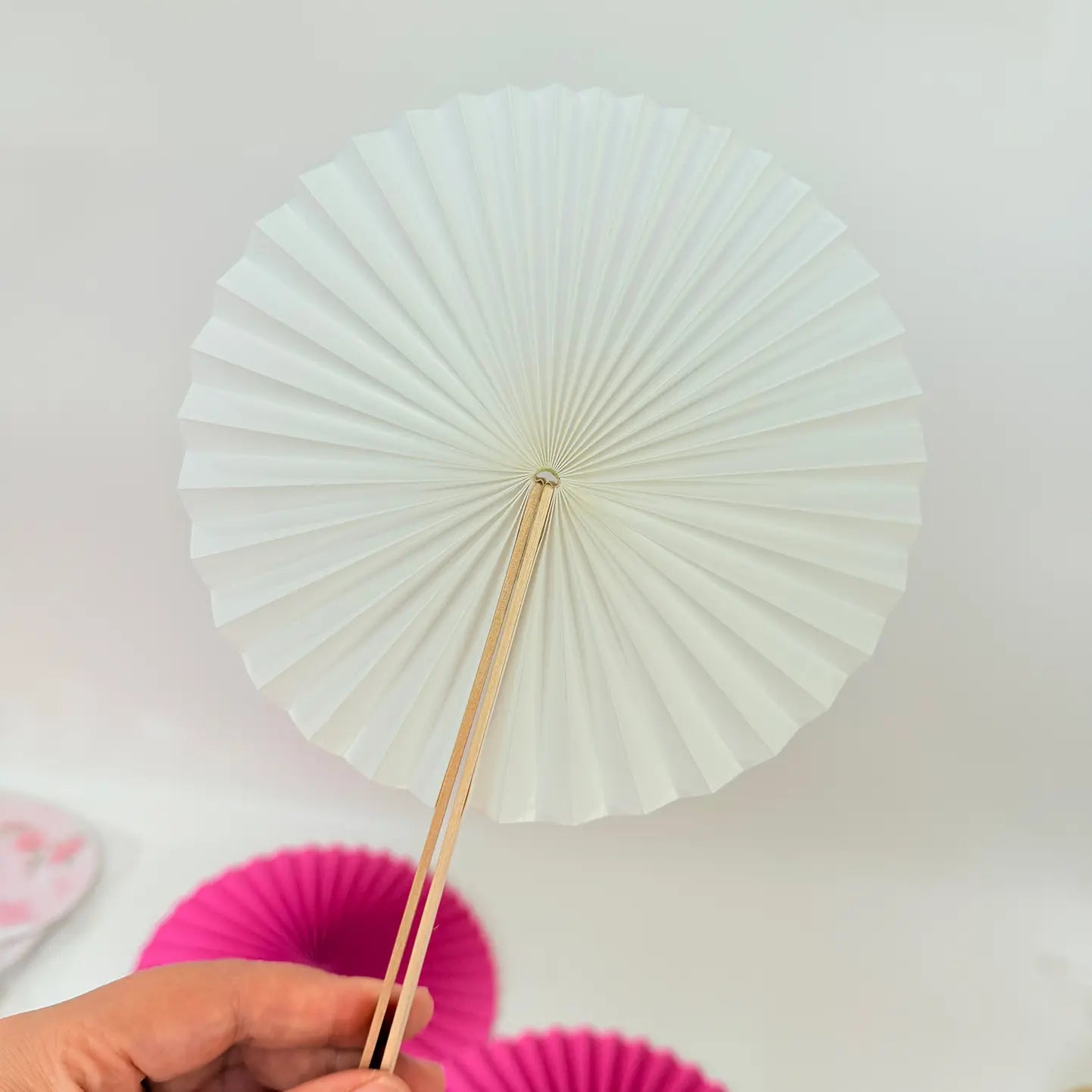A hand holds a white round paper folding fan, with a colorful assortment of other round folding fans featuring diverse designs displayed in the background.