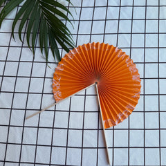 A orange round folding fan lies open on a textured cloth, placed in its half unfolded position. Delicate green leaves are visible in the background.