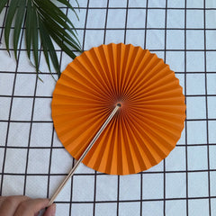 A orange round folding fan lies open on a textured cloth, secured in its unfolded position by a hand. Delicate green leaves are visible in the background.