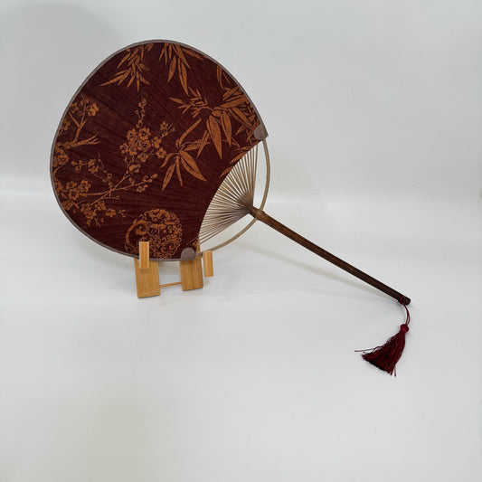 A Japanese round fan is placed on a fan stand. The fan is made of Xiangyunsha silk and has plum blossom and bamboo leaf patterns.