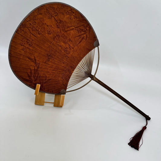 A Japanese round fan is placed on a fan stand. The fan is made of Xiangyunsha silk and has plum blossom and bamboo leaf patterns.