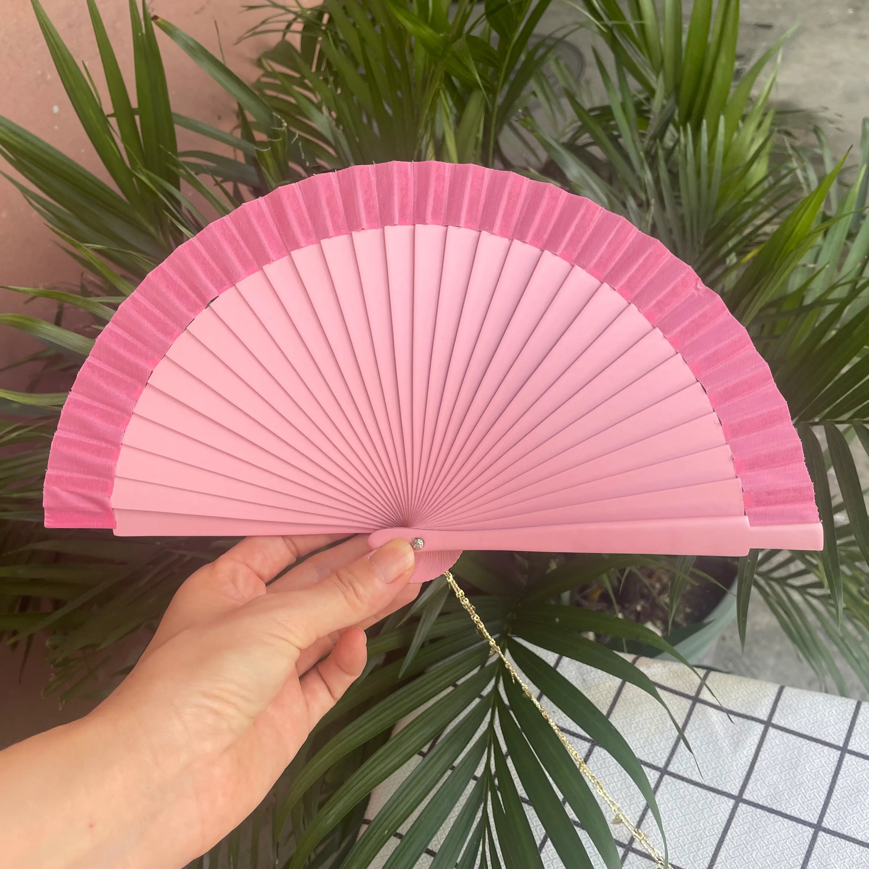 Hand holding a pink Spanish-style fan with a gold charm on the handle. Styled with greenery and white cloth in the background.