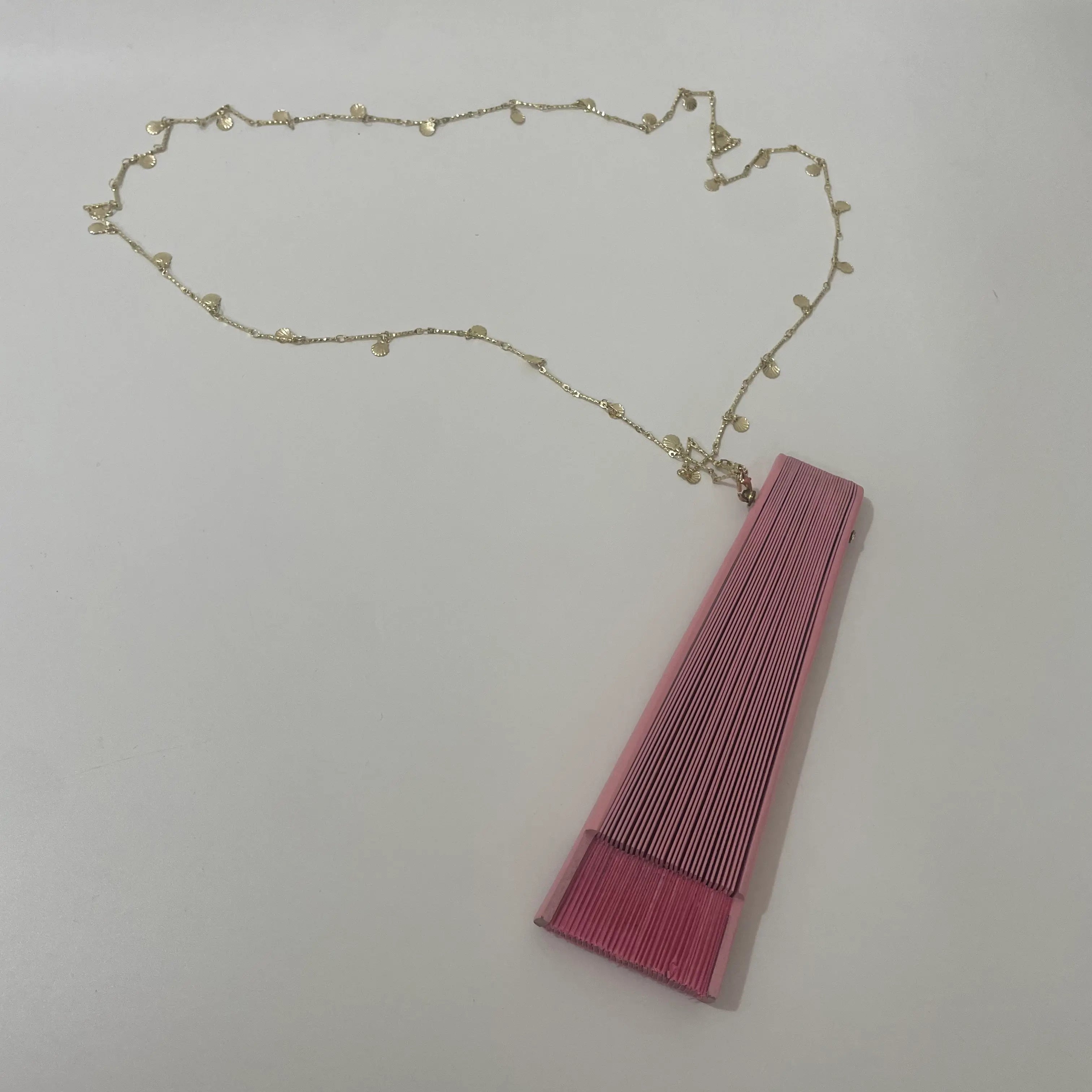 A folded Spanish-style fan in pink with a gold charm on its handle, resting on a white surface.