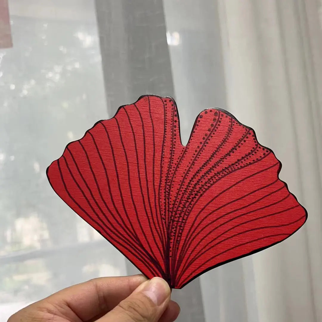 A hand holding a red gingko leaf shape fan panel in front of a white curtain
