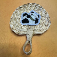 A hand size palm leaf fan on a table, a panda shape sticker is on top of the fan