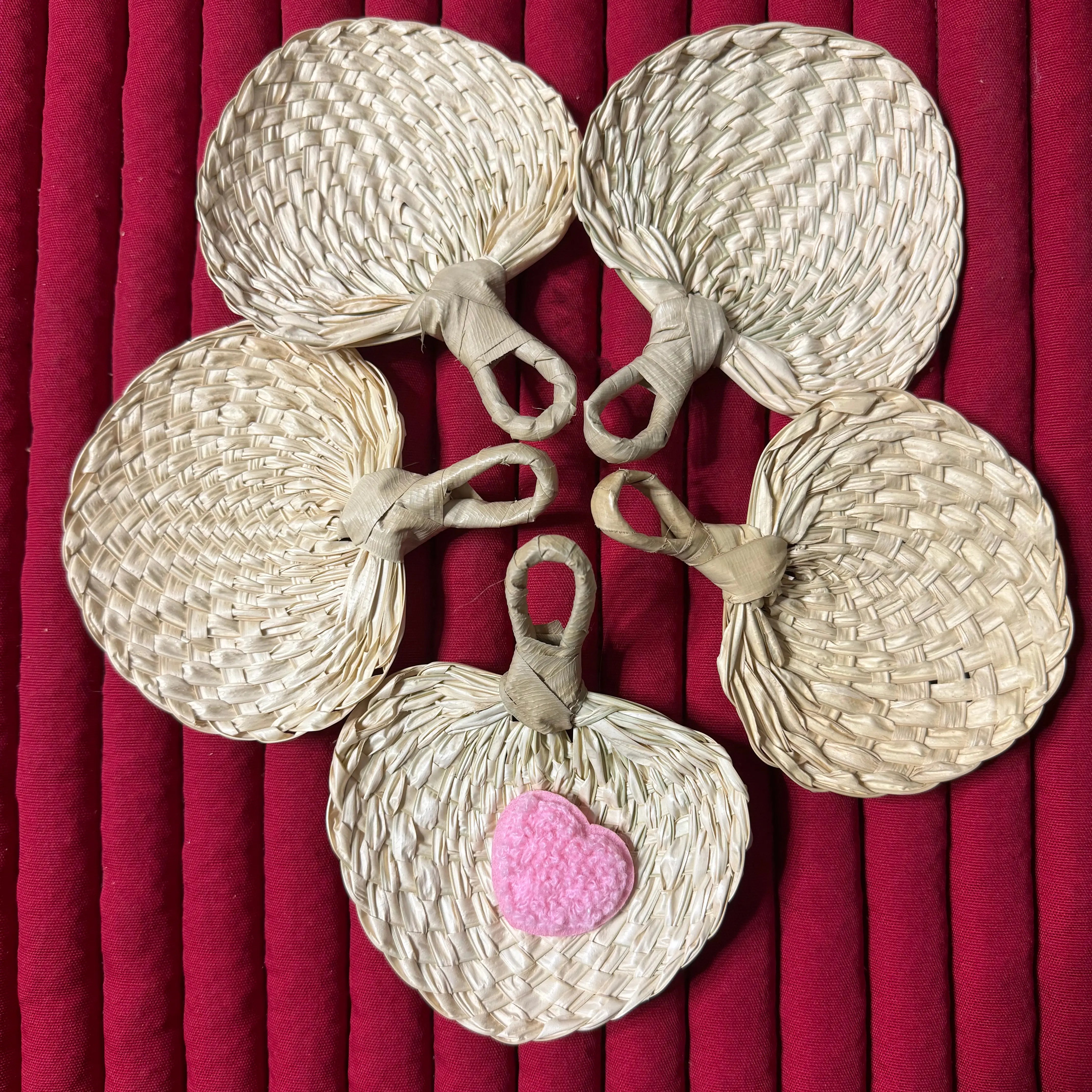 Set of 5 small palm leaf fans arranged on a red cloth. One fan features a distinctive pink heart-shaped decoration.